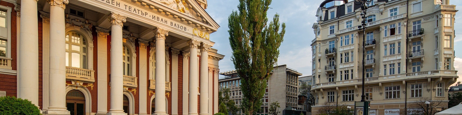 Um teatro imponente com colunas e estátuas clássicas, rodeado por uma praça com piso padronizado e edifícios modernos.