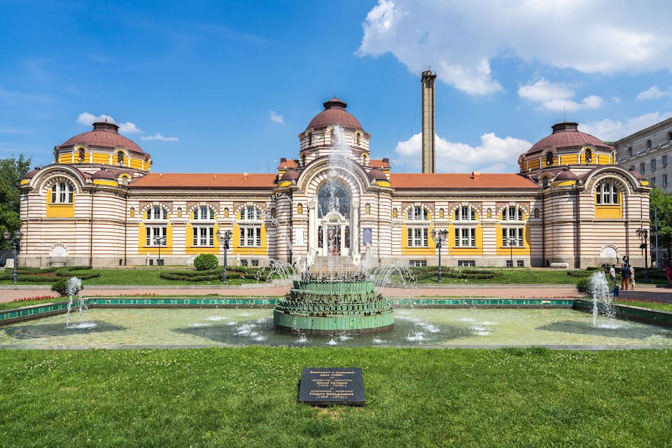 Een historisch gebouw met een centrale fontein, omgeven door een verzorgd gazon en een helderblauwe lucht.