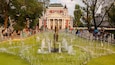 Une grande fontaine ornée d’une statue au centre, entourée d’une foule de personnes. Au fond, on aperçoit un grand bâtiment rouge, doté de colonnes et surmonté d’une statue.