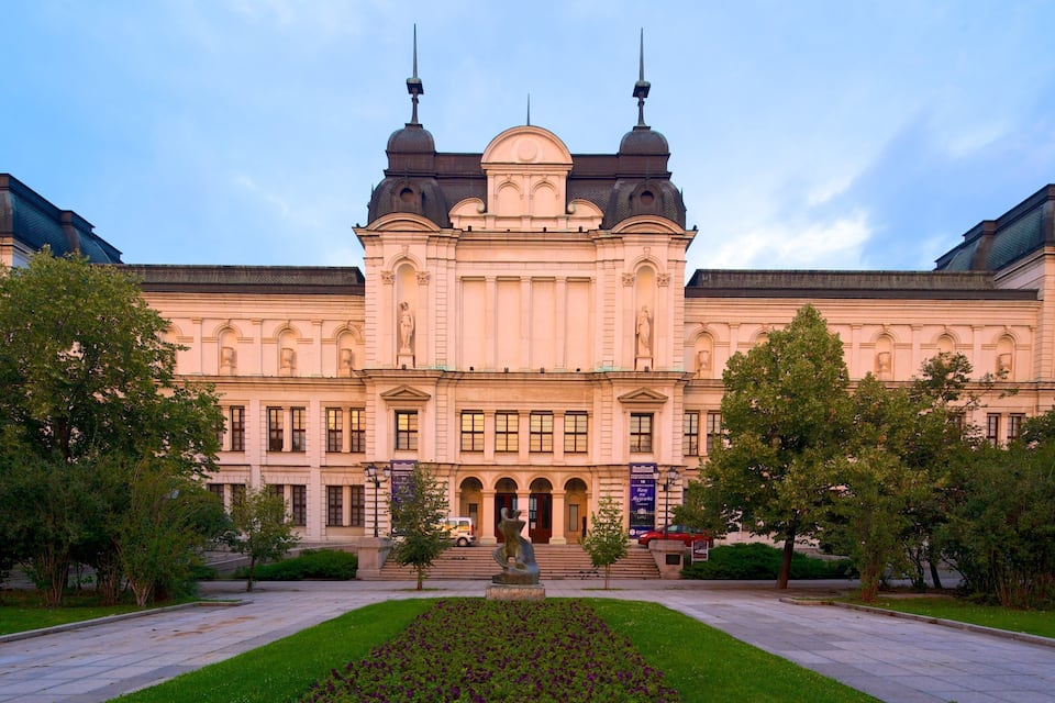 A large, classical building with a central dome and two towers, surrounded by a well-maintained garden and a paved walkway.