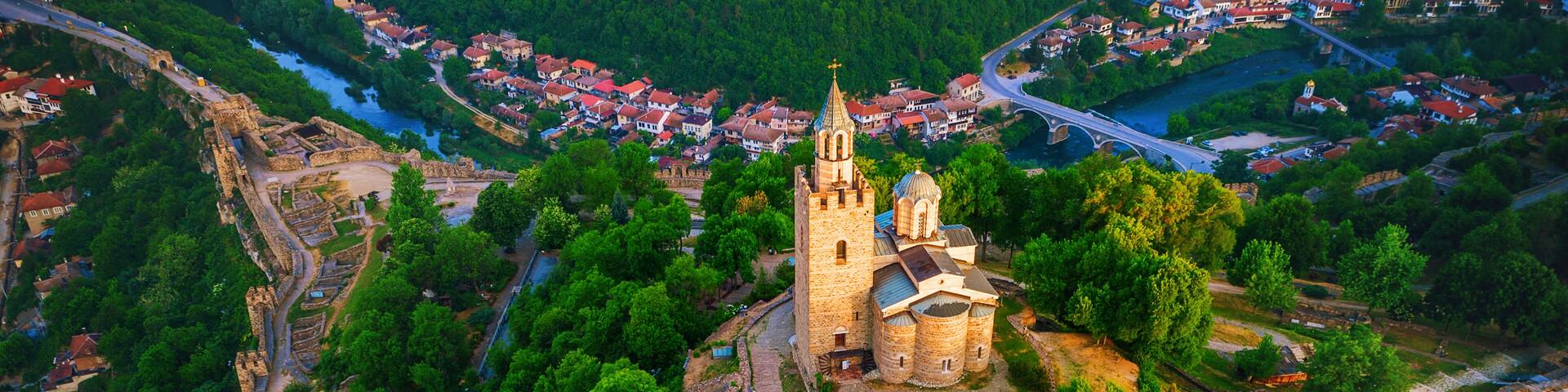 An aerial view of a historic church surrounded by dense forest, a river, and a bridge connecting to a nearby town.