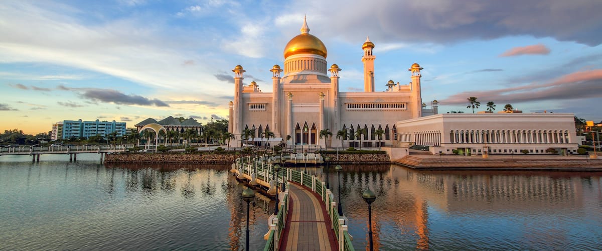 A grand mosque with a golden dome, a reflection in the water, and a bridge leading to it.