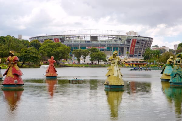 A park with statues of figures in colorful costumes on a lake, a large stadium in the background, and trees around.