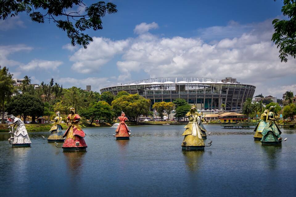 Um lago com estátuas de figuras em trajes coloridos, um estádio moderno ao fundo e árvores ao redor.