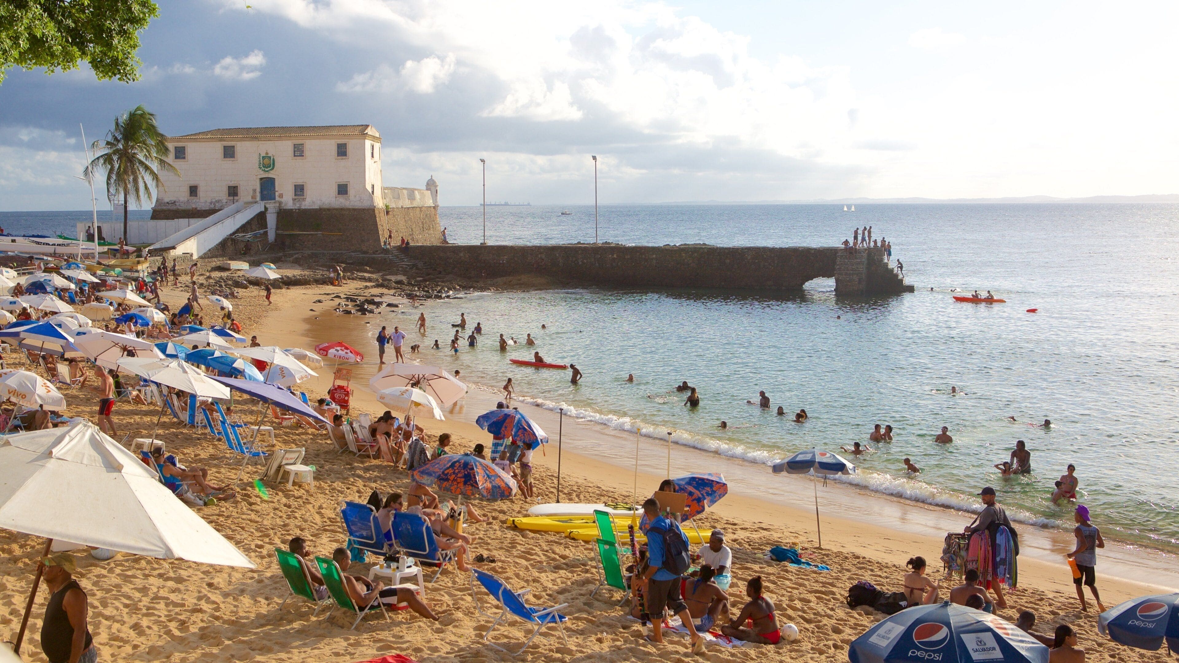 Une plage bondée, avec des parasols, des gens dans l’eau et un bâtiment en arrière-plan.