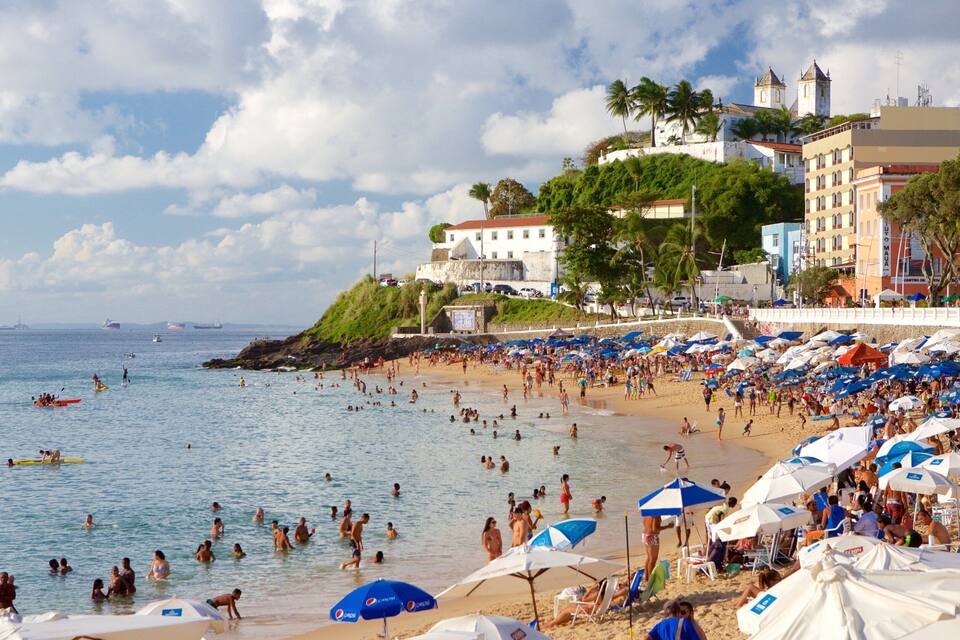 A crowded beach with people swimming and sunbathing, a large building with a tower, and palm trees.