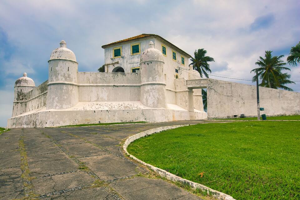 A historic fort with white walls and domed towers, surrounded by a grassy area and palm trees.