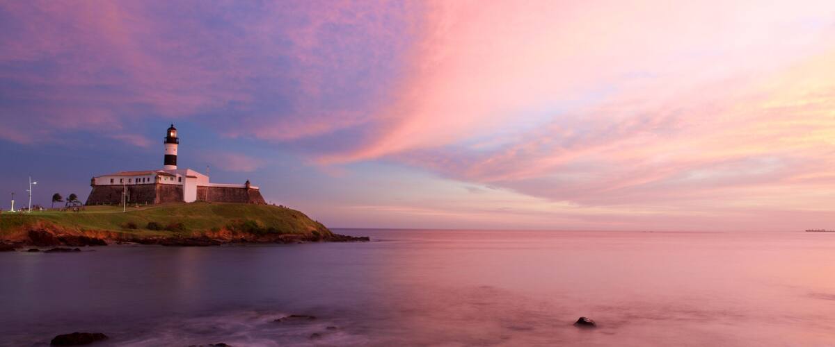 A lighthouse on a grassy hill by the sea with a pink and purple sky.