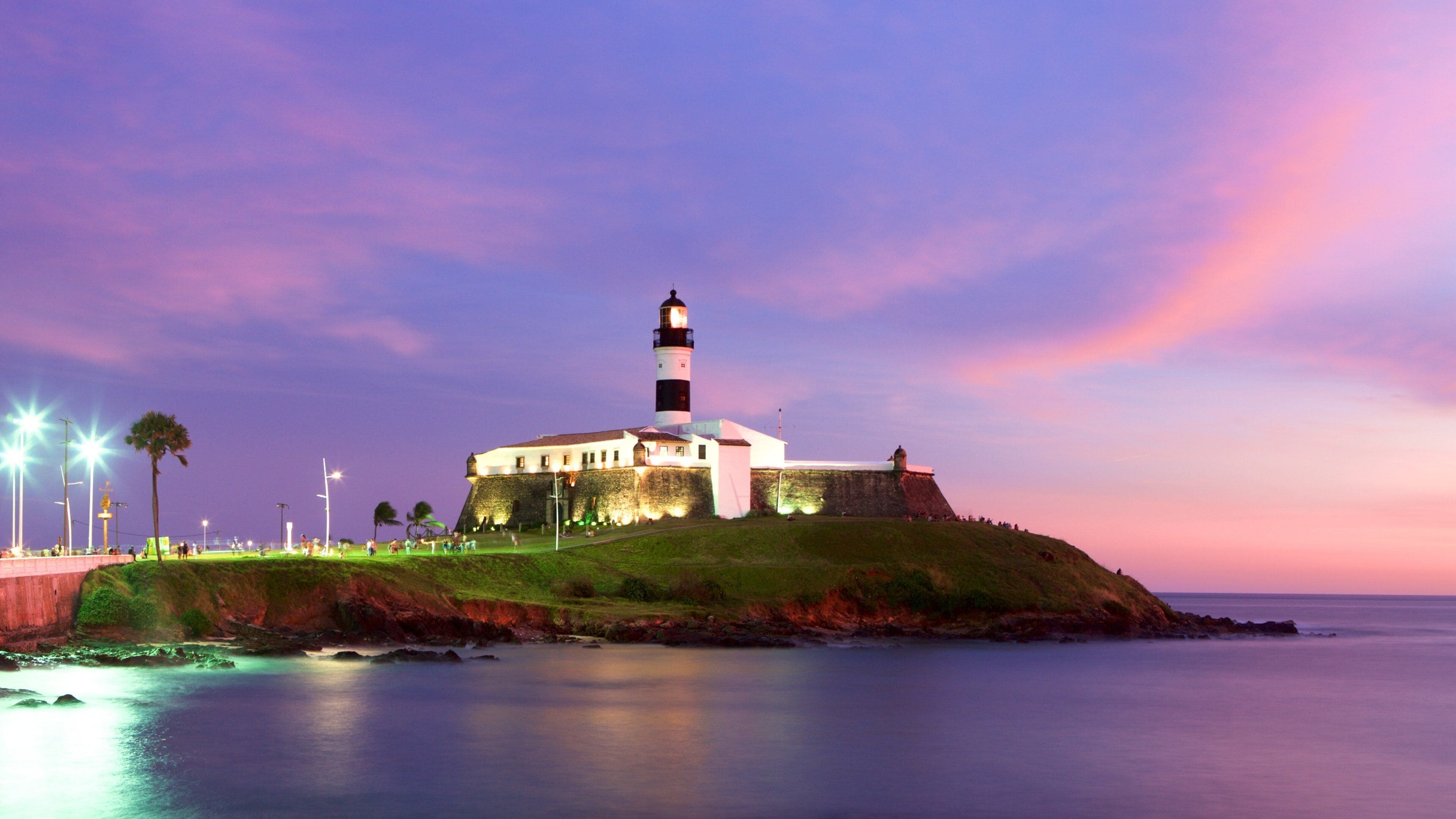 A lighthouse on a grassy hill by the sea, with a colorful sky in the background.