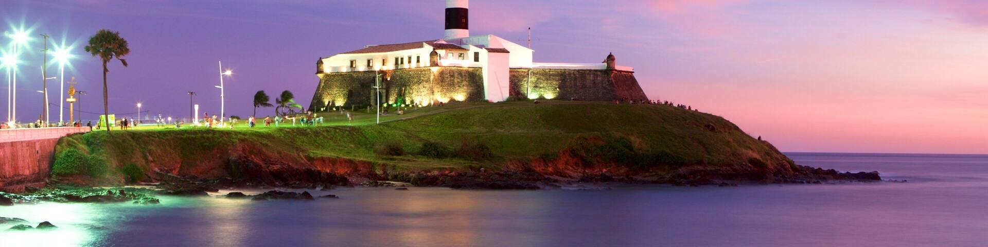 A lighthouse on a grassy hill by the sea, with a colorful sky in the background.