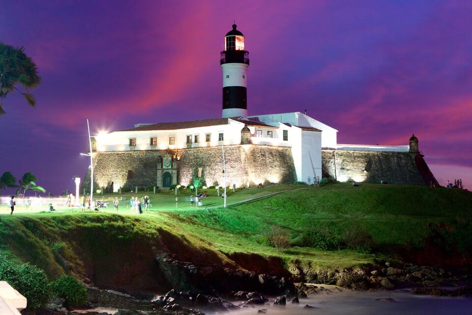 A historic fort with a lighthouse, surrounded by a grassy hill and a beach.
