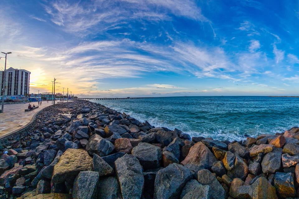 Una costa rocosa con un muelle que se adentra en el mar, un cielo despejado y un paisaje urbano al fondo.