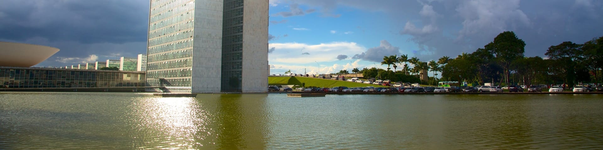 Un edificio moderno y alto con fachada de vidrio, una masa de agua al frente y un cielo parcialmente nublado.