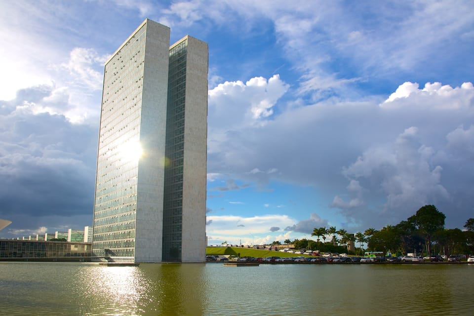 Um edifício alto e moderno com fachada de vidro, um corpo de água em primeiro plano e um céu parcialmente nublado.