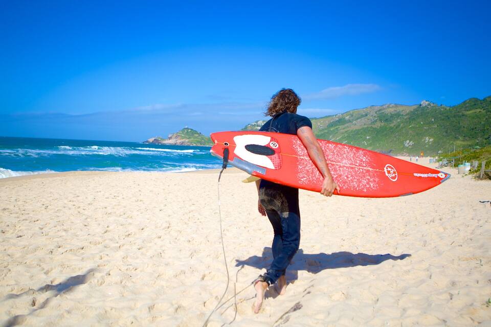 A person carrying a red surfboard on a sandy beach with waves in the background and hills in the distance.