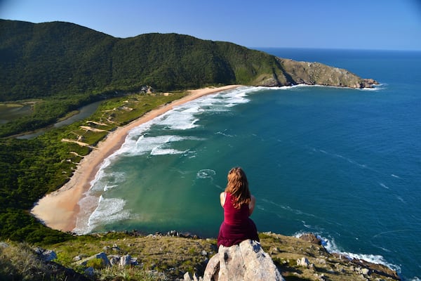 Une femme contemple un paysage cĂŽtier comprenant une plage, lâocĂ©an et des collines boisĂ©es.
