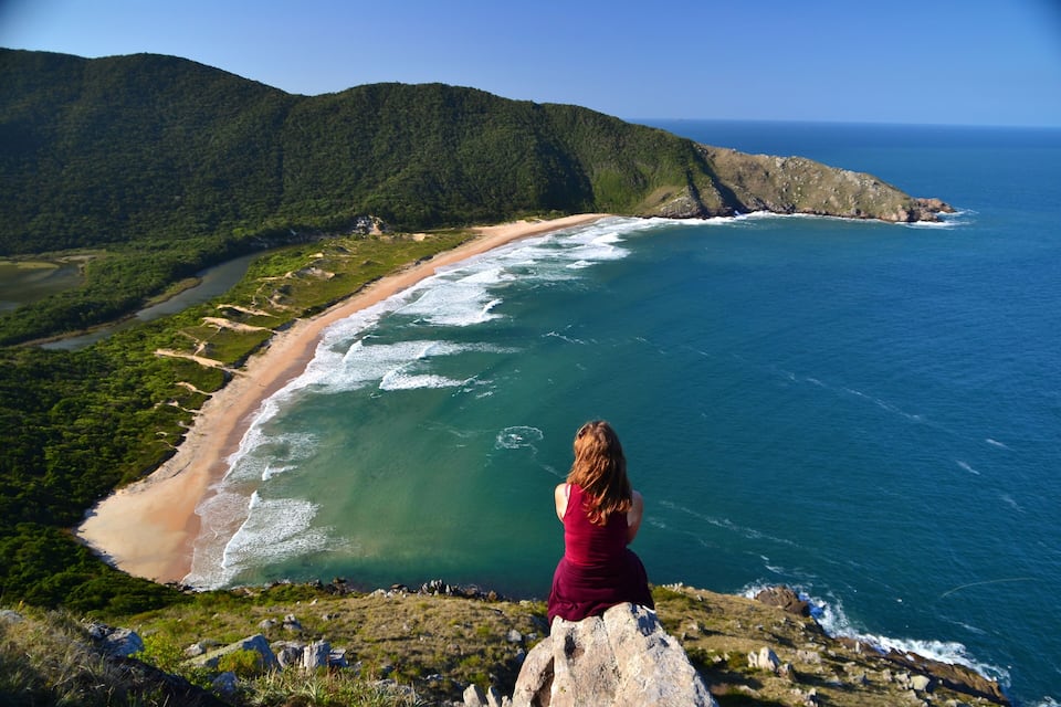 Una mujer observa un paisaje costero con playa, océano y colinas boscosas.