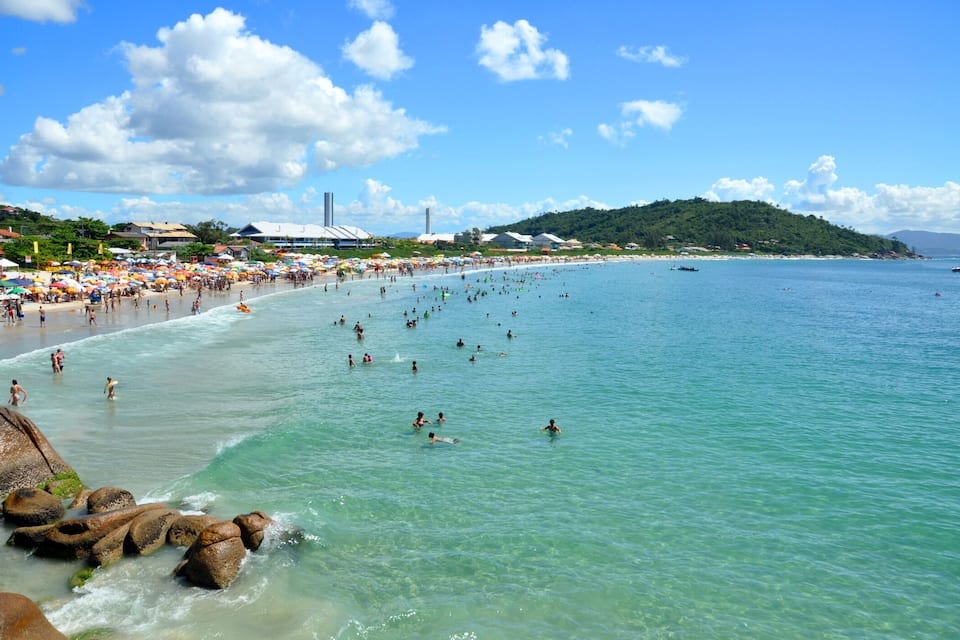 Una playa concurrida, con agua turquesa cristalina, formaciones rocosas y una colina distante con edificios.