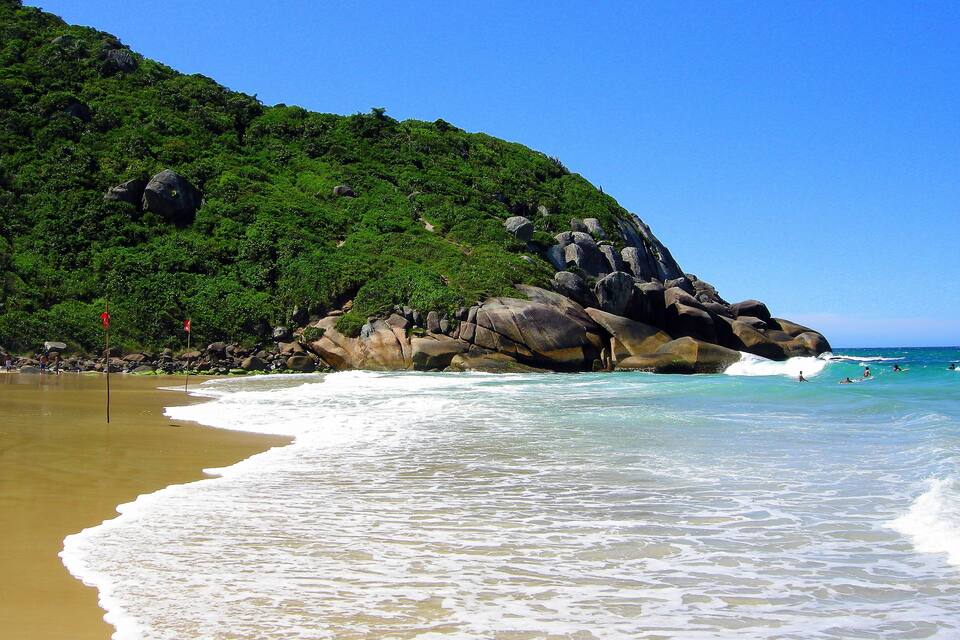 A beach with clear turquoise water, a sandy shore, and a green hill with rocks.