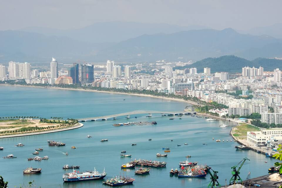 A coastal cityscape with a marina filled with boats, a bridge, and modern buildings.
