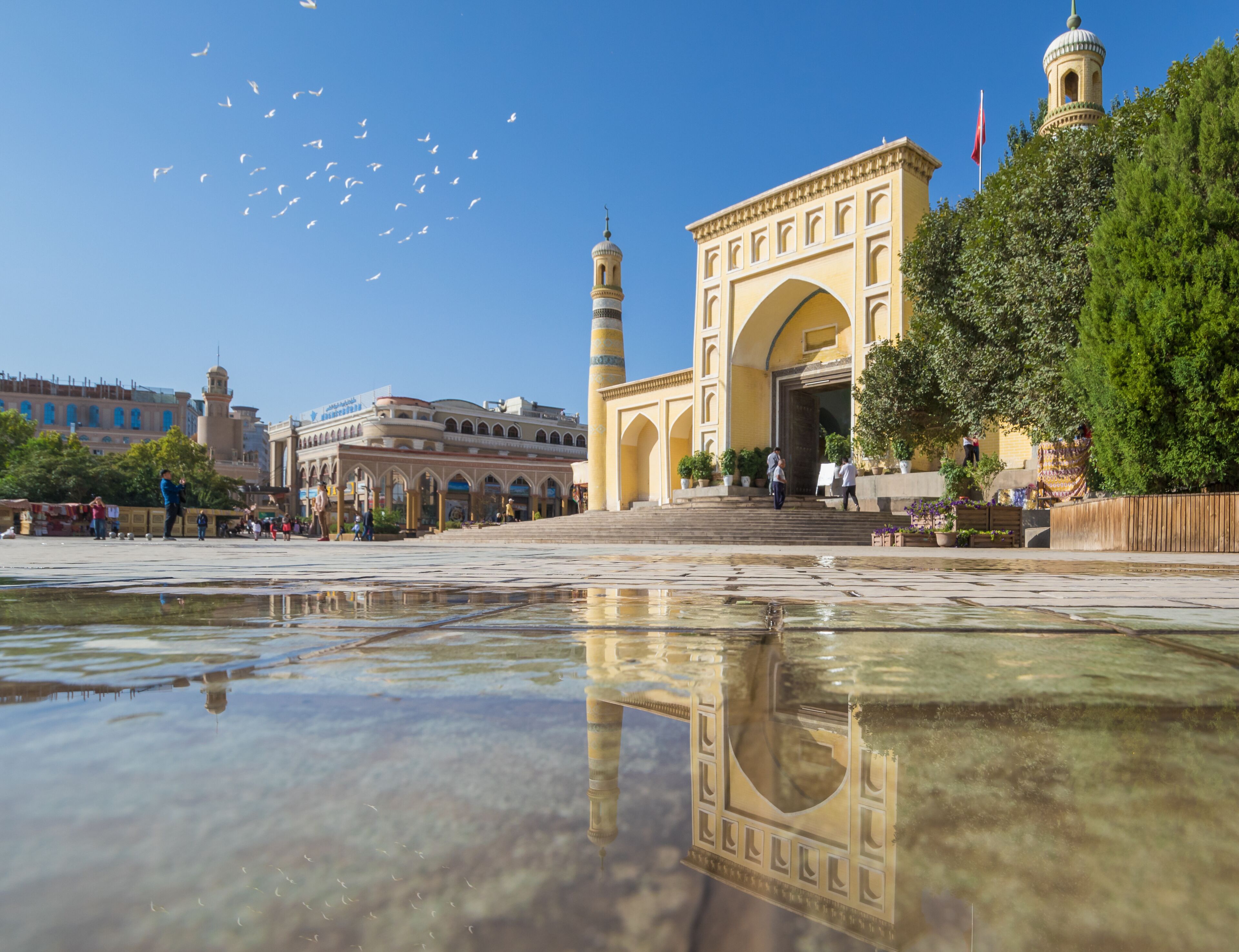 A historic building with an archway and a minaret, surrounded by a reflective water surface and greenery.