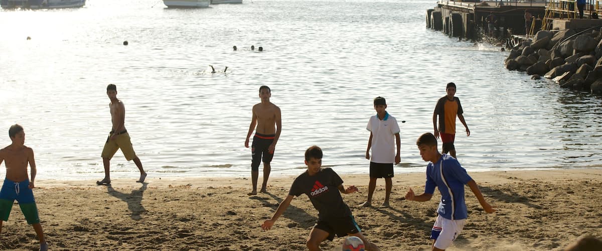 A group of people playing soccer on a sandy beach near a body of water with boats in the background.