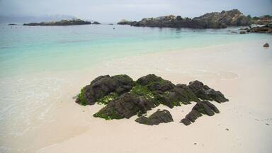 A beach with clear turquoise water, a rocky outcrop covered in green algae, and distant rocky islands.