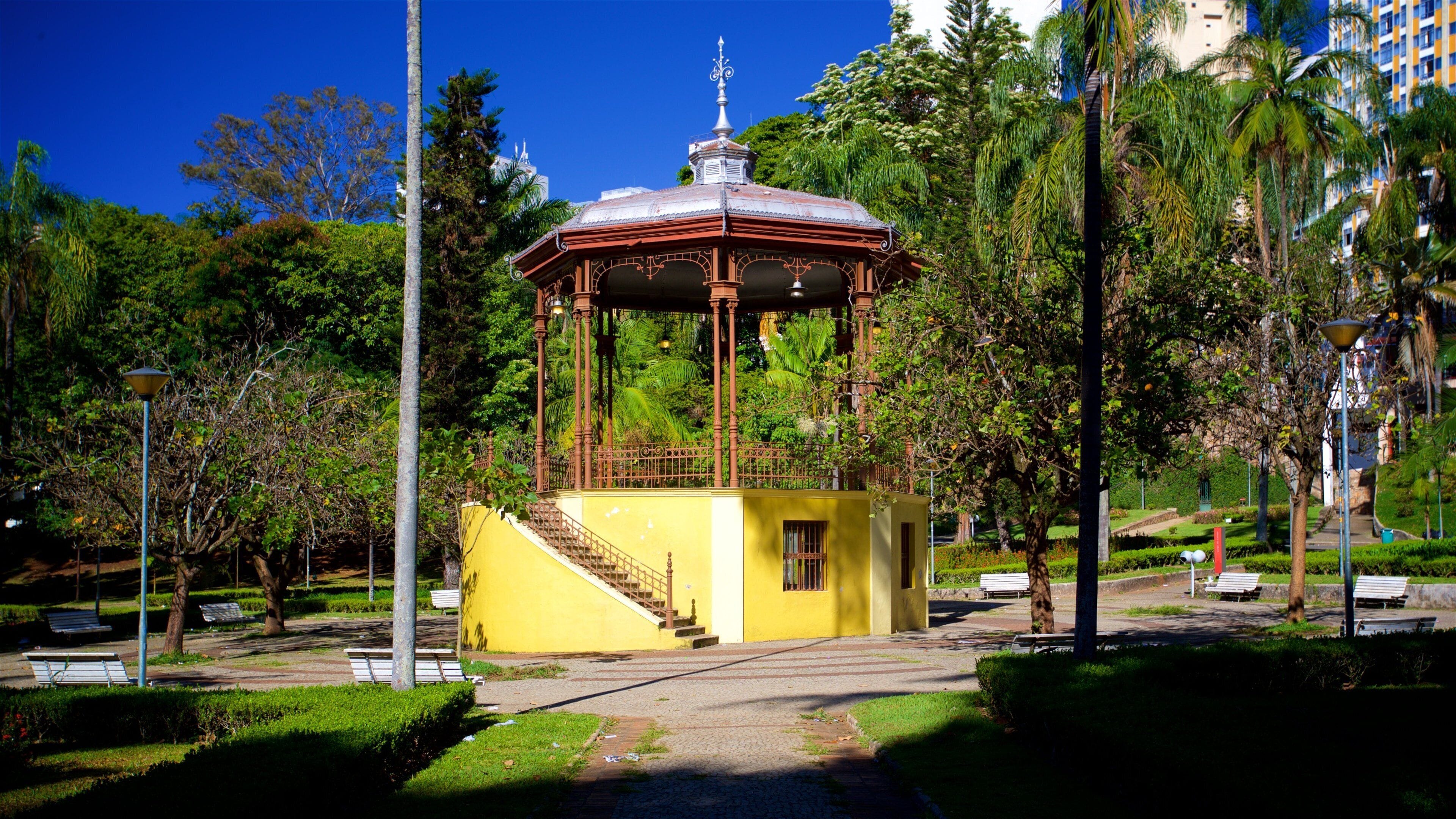 Un kiosque jaune avec un toit en métal et un escalier menant à sa plateforme, entouré d’arbres et de bancs dans un parc.