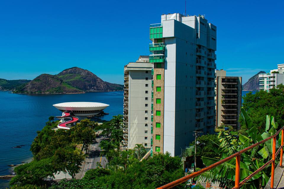 Paisagem costeira com um edifício moderno em destaque, uma vasta extensão de água e montanhas ao fundo.
