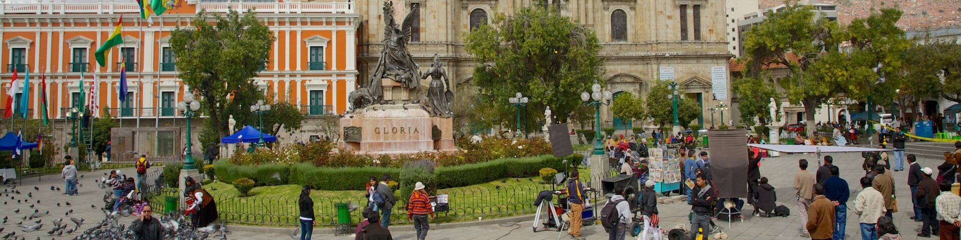 A plaza with a large building featuring domed architecture, a statue, and a fountain.