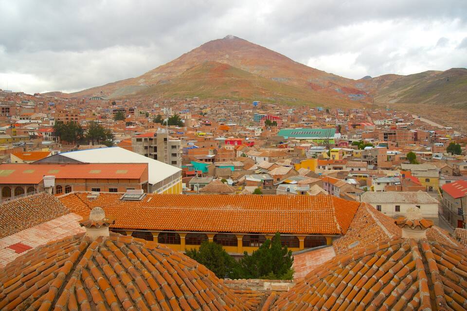 A cityscape with terracotta-roofed buildings, a prominent mountain in the background, and a cloudy sky.