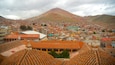 A cityscape with terracotta-roofed buildings, a prominent mountain in the background, and a cloudy sky.