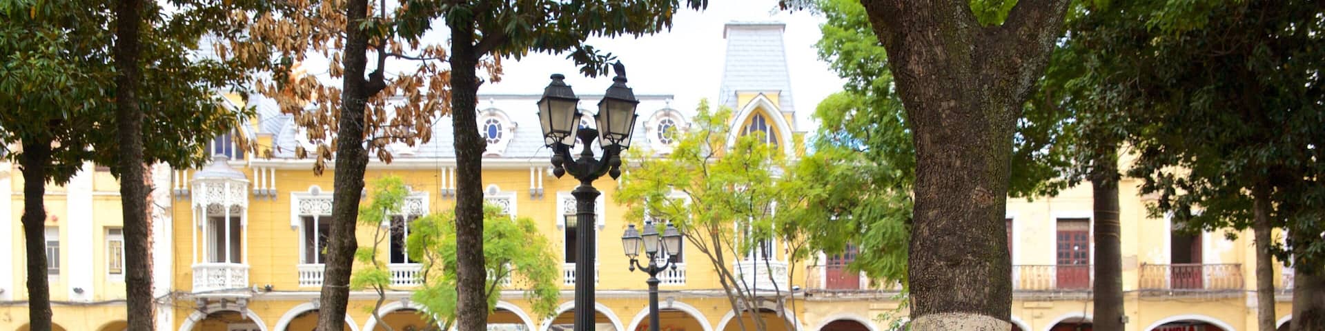Un parque con parterres, árboles y un edificio amarillo con arcos al fondo.