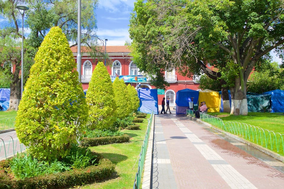 A well-maintained park with neatly trimmed hedges, a paved walkway, and a red building with arched windows in the background.