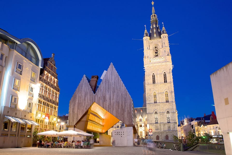 A historic tower with a spire, a modern building with a wooden facade, and a cobblestone square with outdoor seating.