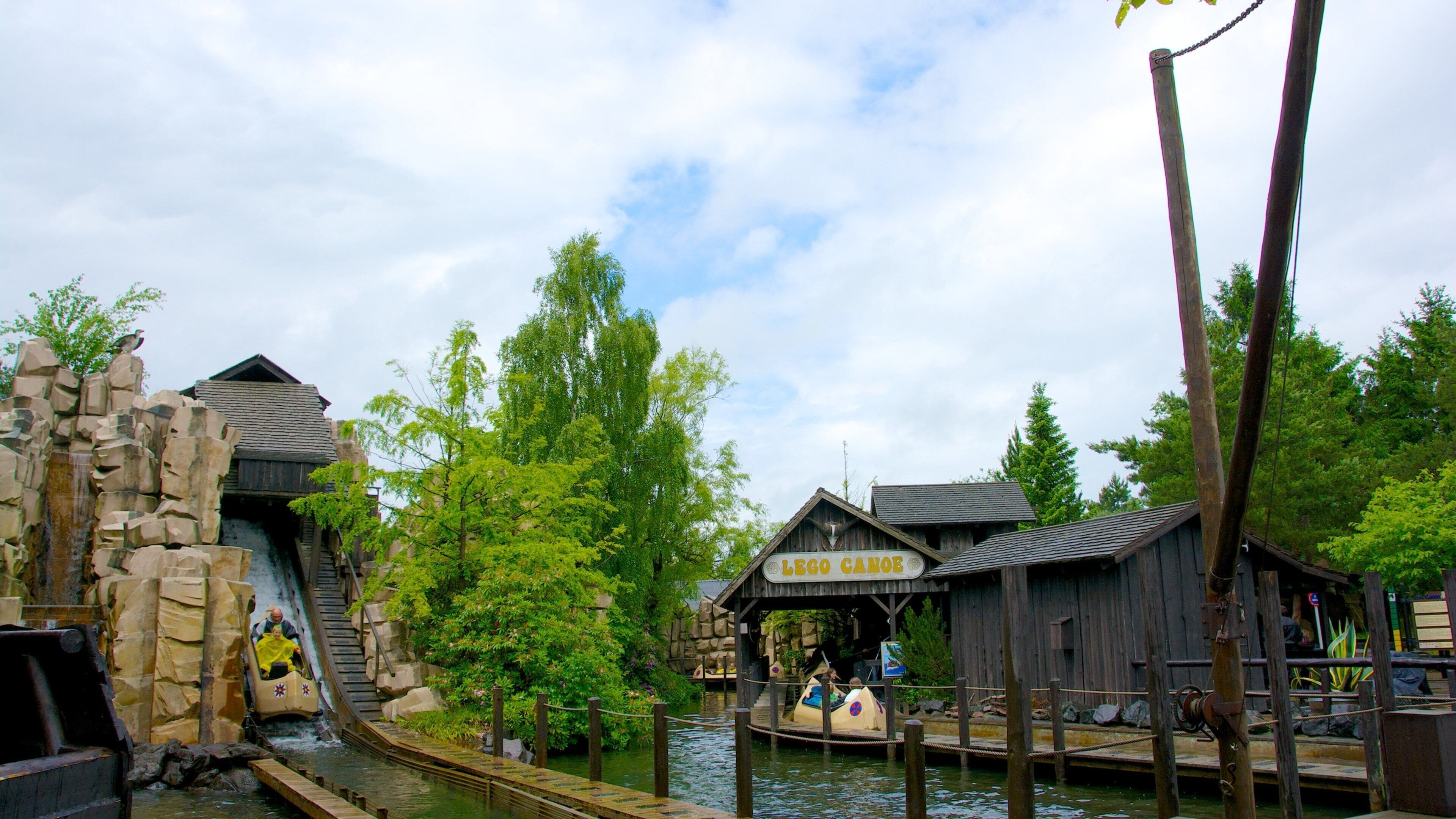A wooden water ride with a LEGO-themed sign, surrounded by greenery and a cloudy sky.