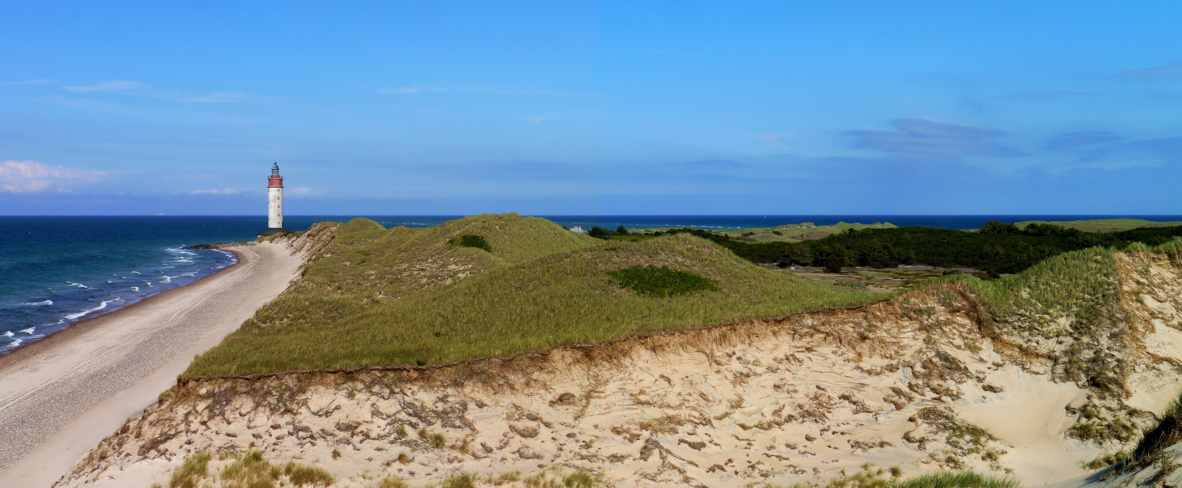 A coastal landscape with a lighthouse, sandy dunes, and the ocean.