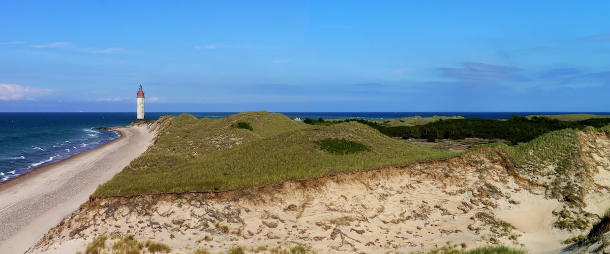 A coastal landscape with a lighthouse, sandy dunes, and the ocean.