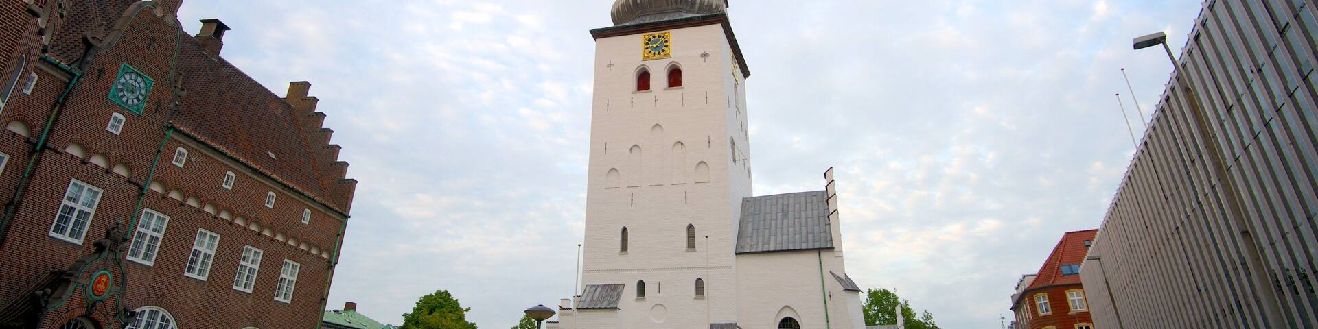 A historic church with a tall bell tower, surrounded by modern buildings and parked cars.
