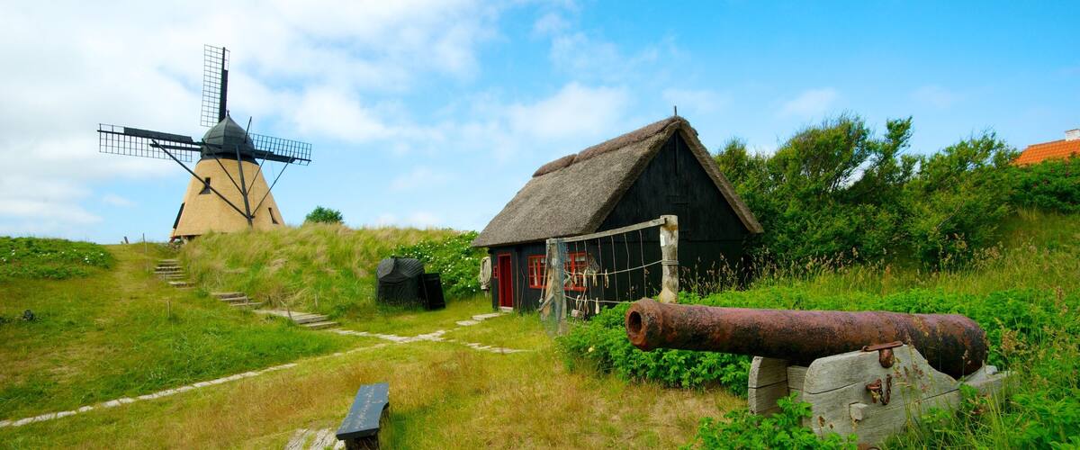 A traditional windmill, a thatched-roof house, and a rusty cannon on a grassy hill.