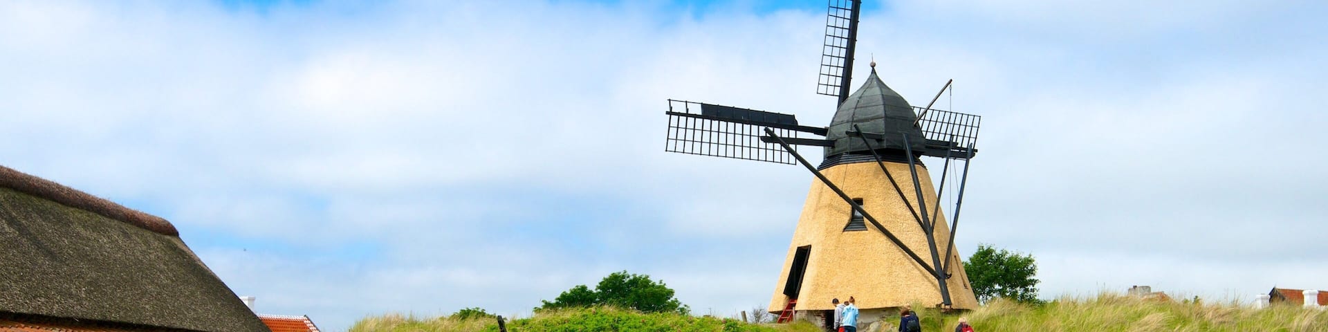 A traditional windmill on a grassy hill with a thatched-roof building nearby.