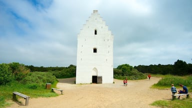 A white tower with a pointed roof, surrounded by greenery and a gravel path.