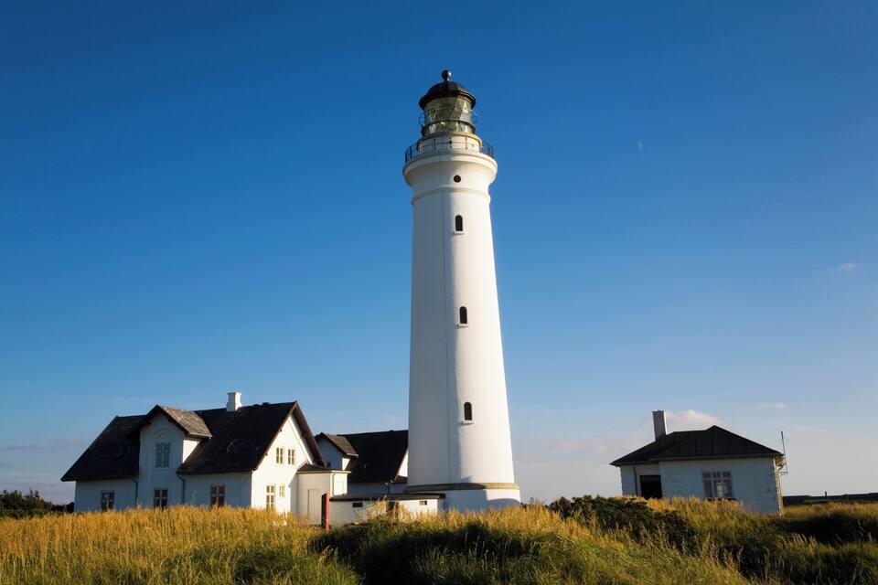Un faro blanco con la parte superior negra, rodeado por dos casas blancas con techos oscuros.