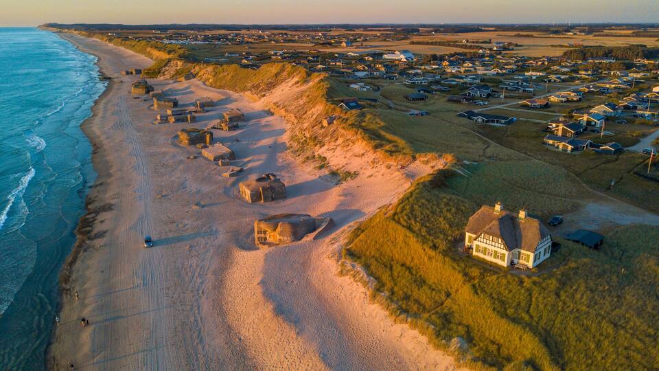 Uma vila costeira com casas sobre dunas, uma praia e campos ao fundo.