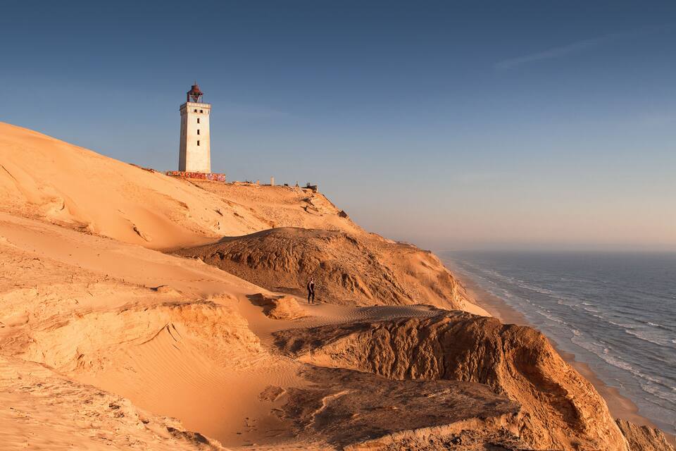 Paesaggio costiero con un faro su una scogliera sabbiosa che si affaccia sul mare.