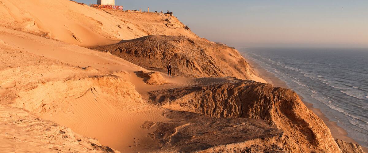 A coastal landscape with a lighthouse on a sandy cliff overlooking the sea.