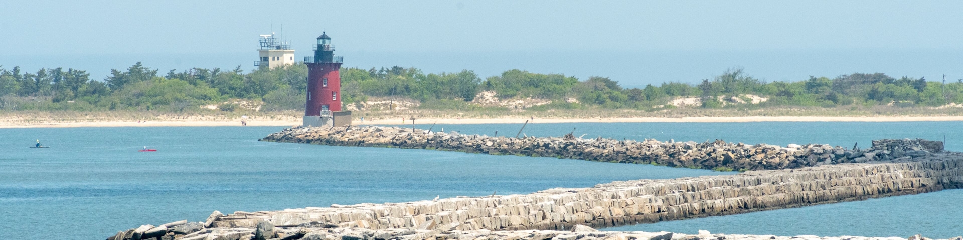 Um farol, um molhe rochoso, mar calmo e uma praia distante com vegetação.