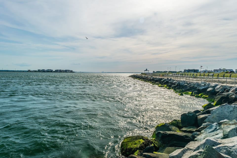 A coastal scene with a rocky breakwater, calm water, and distant buildings.