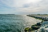 A coastal scene with a rocky breakwater, calm water, and distant buildings.