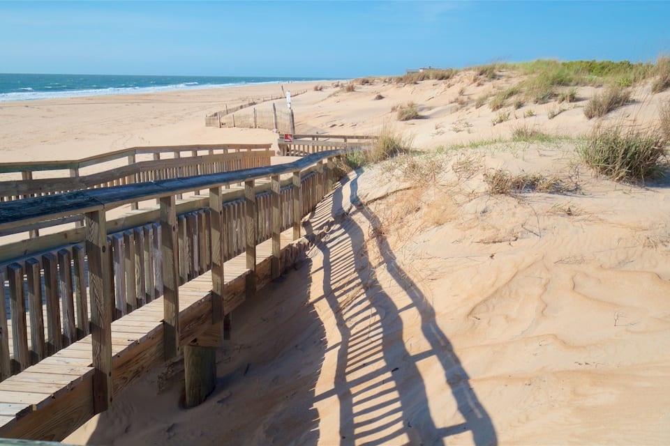 A wooden boardwalk leading to a sandy beach with dunes in the background.